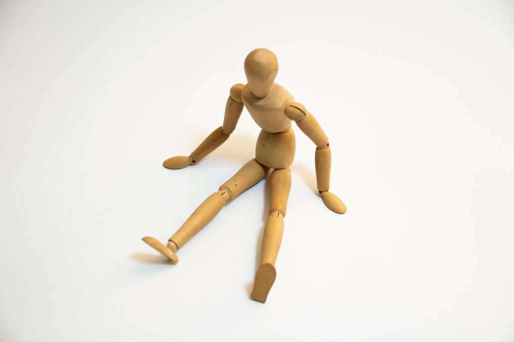 Eine am Boden sitzende, kraftlos nach vorne gebeugte Gliederpuppe aus Holz als Symbol für Immobilität, Bettlägerigkeit und den hohen Unterstützungsbedarf bei Pflegegrad 4