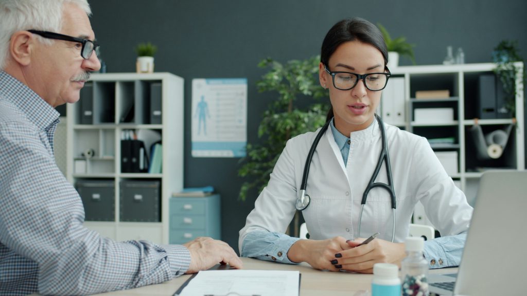 Eine Ärztin mit Brille und Stethoskop berät einen älteren Mann am Schreibtisch über die medizinischen Voraussetzungen für die Berechnung des Pflegegrads
