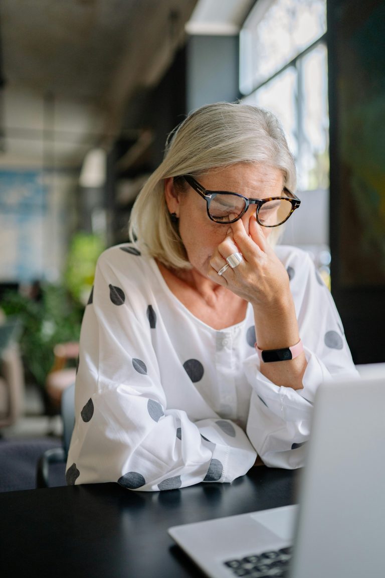 Eine reifere Frau mit Brille sitzt gestresst und erschöpft vor ihrem Laptop und fasst sich an die Nasenwurzel, als Symbol für Überforderung bei der Berechnung des Pflegegrads