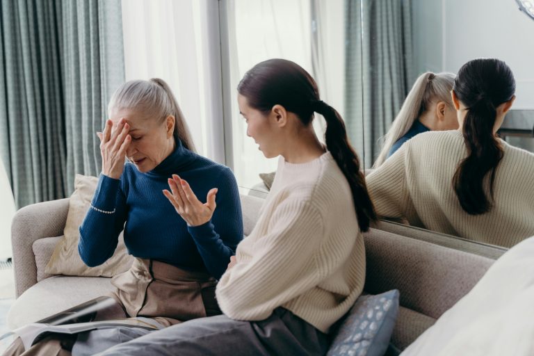 Eine verzweifelte ältere Frau hält sich auf dem Sofa sitzend die Hand vor den Kopf, während eine jüngere Angehörige besorgt mit ihr spricht, als Symbol für psychische Überforderung und Probleme beim Pflegegrad-Antrag