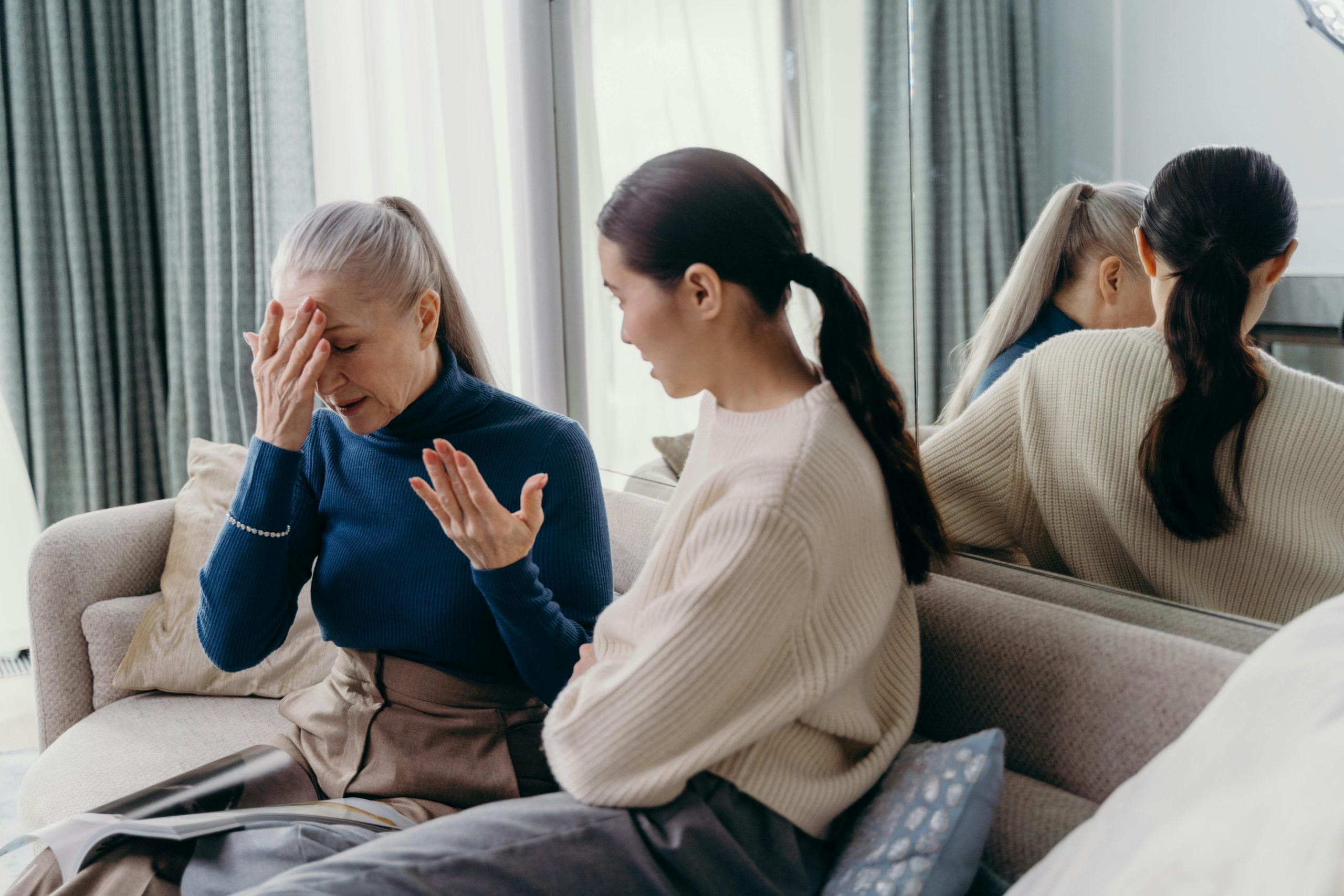 Eine verzweifelte ältere Frau hält sich auf dem Sofa sitzend die Hand vor den Kopf, während eine jüngere Angehörige besorgt mit ihr spricht, als Symbol für psychische Überforderung und Probleme beim Pflegegrad-Antrag